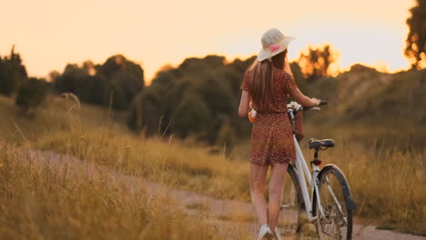 Back-plan-slow-motion:-a-Beautiful-blonde-in-a-dress-with-flowers-in-a-basket-and-a-retro-bike-walks-along-the-road-in-the-summer-field-looking-around-and-smiling-feeling-free.